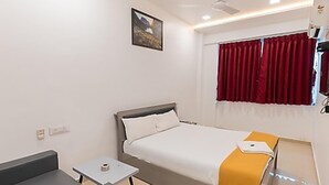 A modern hotel room with a bed, a small table, a chair, a window with red curtains, and a ceiling fan. - Hotel Nirali Rooms - Nexus Hospitality (Surat)