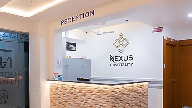 A reception area with a stone-textured counter, a sign reading "RECEPTION," and a logo for "NEXUS HOSPITALITY." - Hotel Nirali Rooms - Nexus Hospitality (Surat)