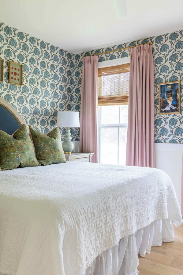 Un dormitorio con una cama grande, papel tapiz floral, una ventana con cortinas y una lámpara de noche.