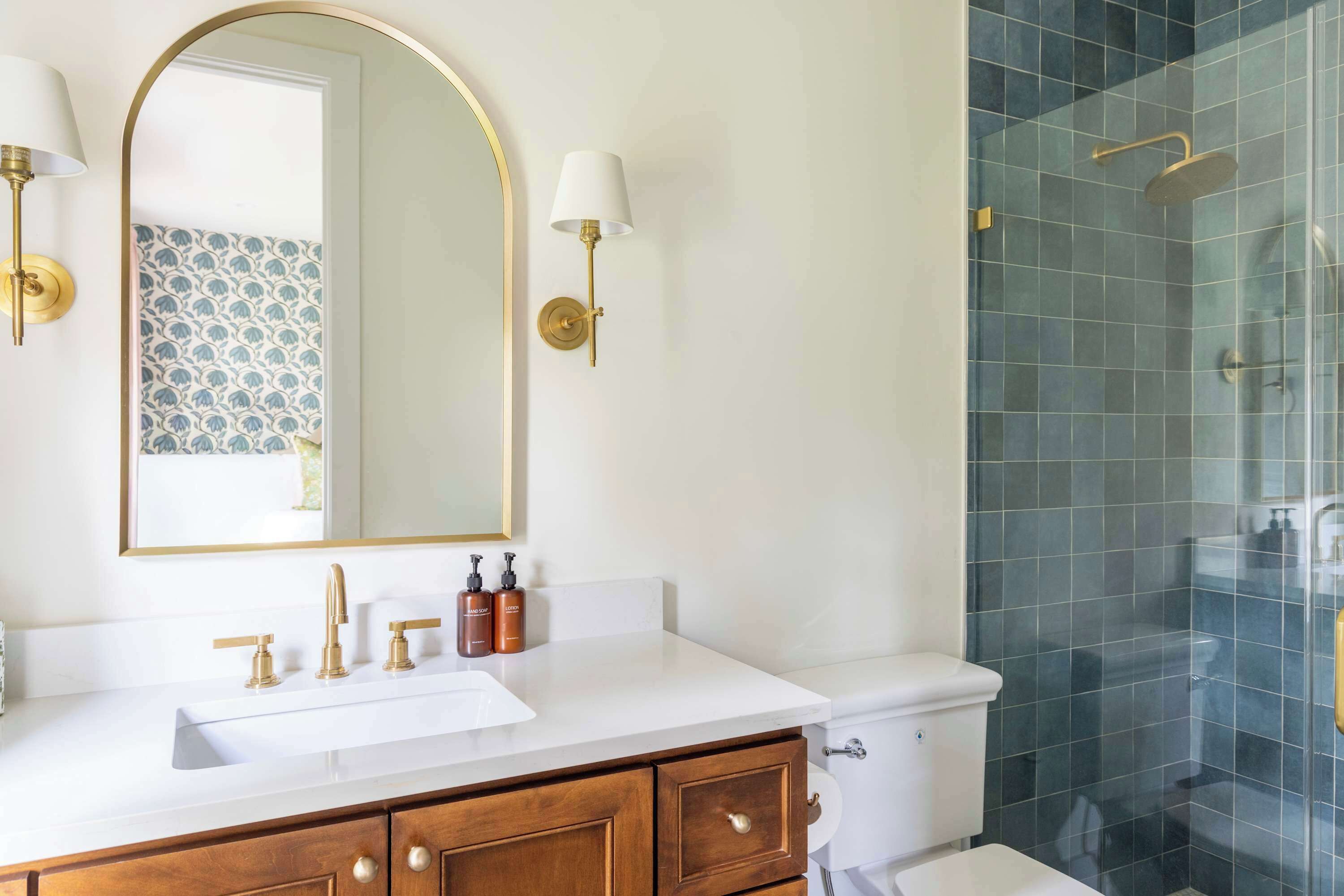 A bathroom with a white sink, gold faucets, a wooden vanity, a large mirror, and a shower with blue tiles.