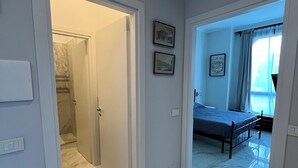 Una habitación de hotel con cama, baño con ducha y pasillo con puerta que da a otra habitación.