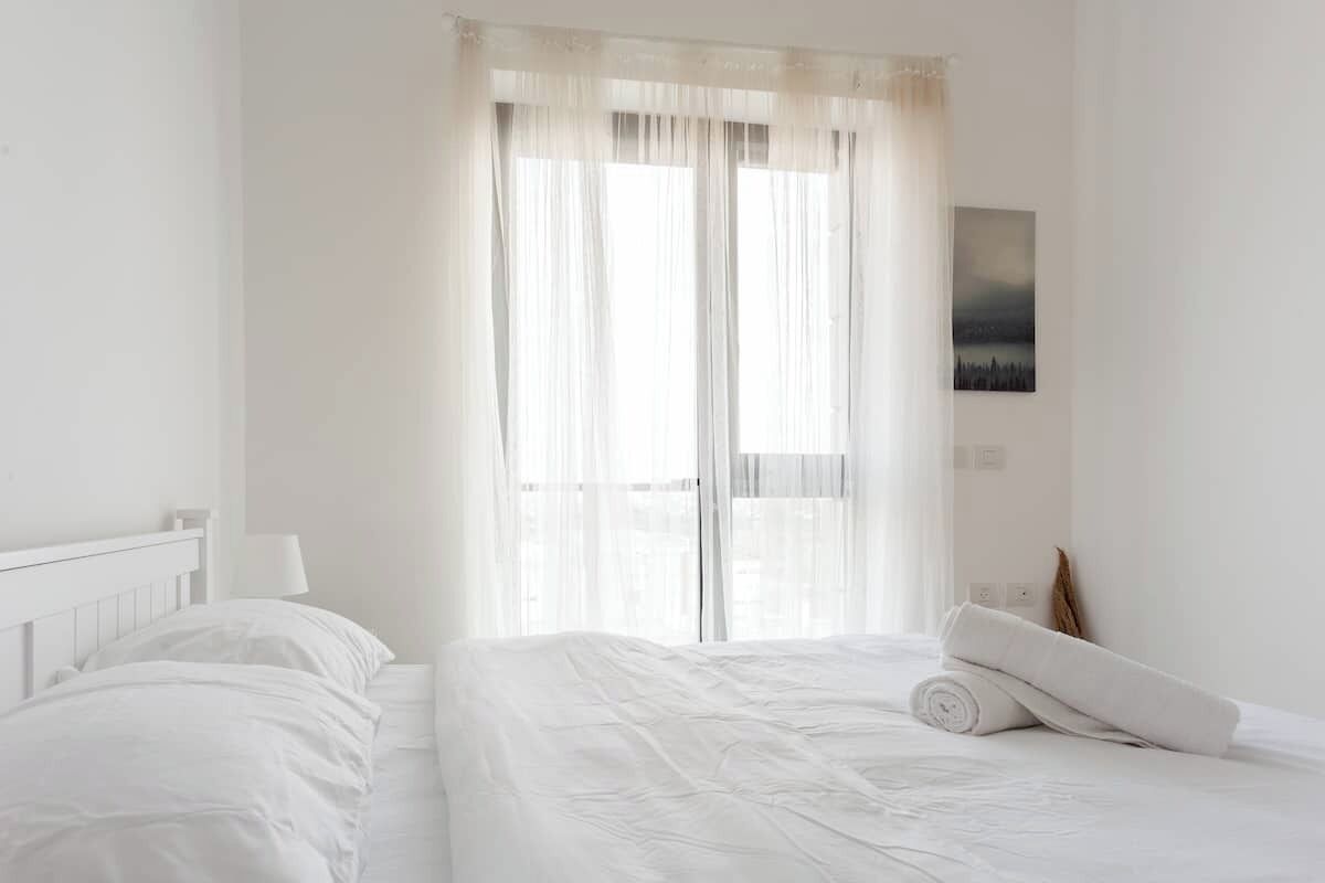 A bedroom with a large bed, a nightstand, a lamp, and a window with sheer curtains.