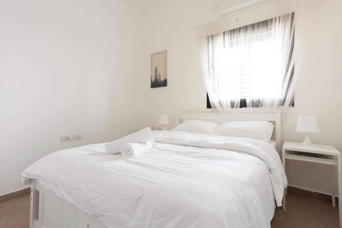 A white bedroom with a bed, a nightstand, a lamp, and a window with curtains.