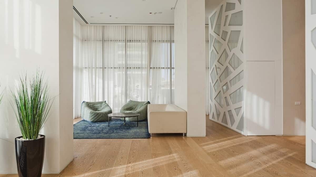 Ein modernes Wohnzimmer mit Holzboden, einem großen Fenster mit durchscheinenden Vorhängen, einem blauen Teppich und einer Sitzecke mit Sofa und Sesseln.