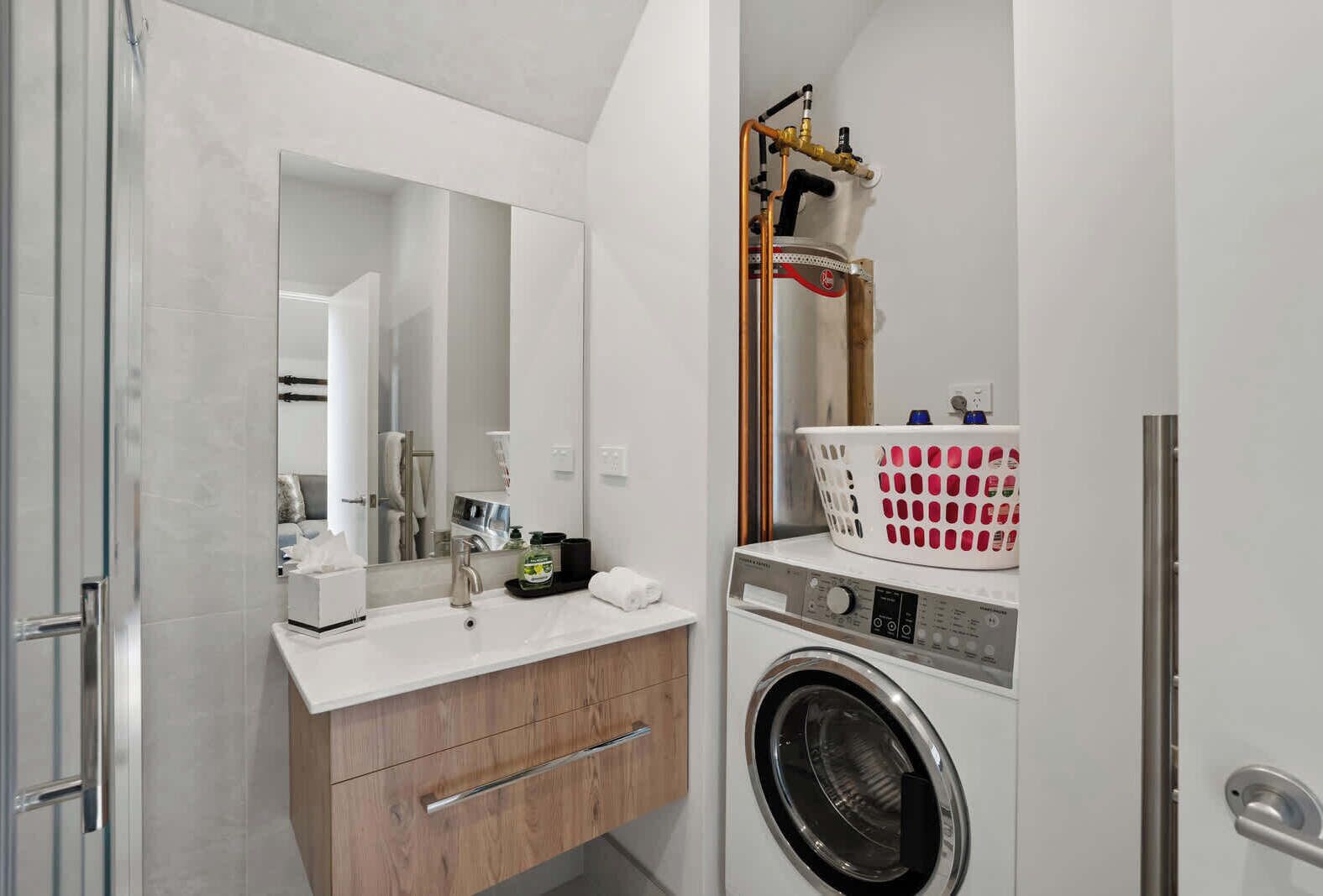 A modern bathroom with a white sink, wooden vanity, a large mirror, a washing machine, and a laundry basket.