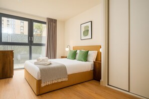 A modern bedroom with a wooden bed, a large window with curtains, a framed picture on the wall, and a wooden nightstand. - Calm and Cozy Corner (London)