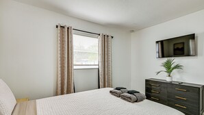 Een slaapkamer met een bed, een dressoir, een aan de muur bevestigde televisie en een raam met gordijnen.