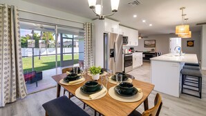 A modern kitchen with a central island, a dining area with a table set for four, and a view of a green lawn through large sliding glass doors. - Game, Shop & Beach-hop: Family Home in Cape Coral (Cape Coral)