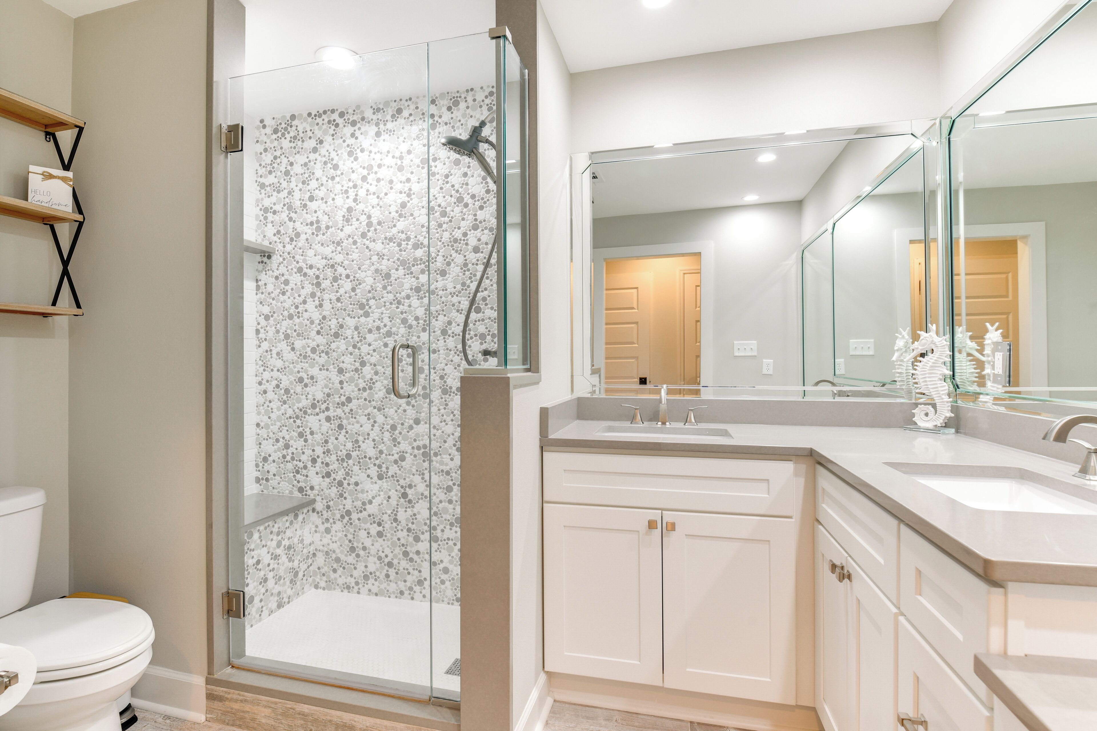 A modern bathroom with a glass-enclosed shower, a white toilet, a vanity with a large mirror, and a shelf with decorative items.