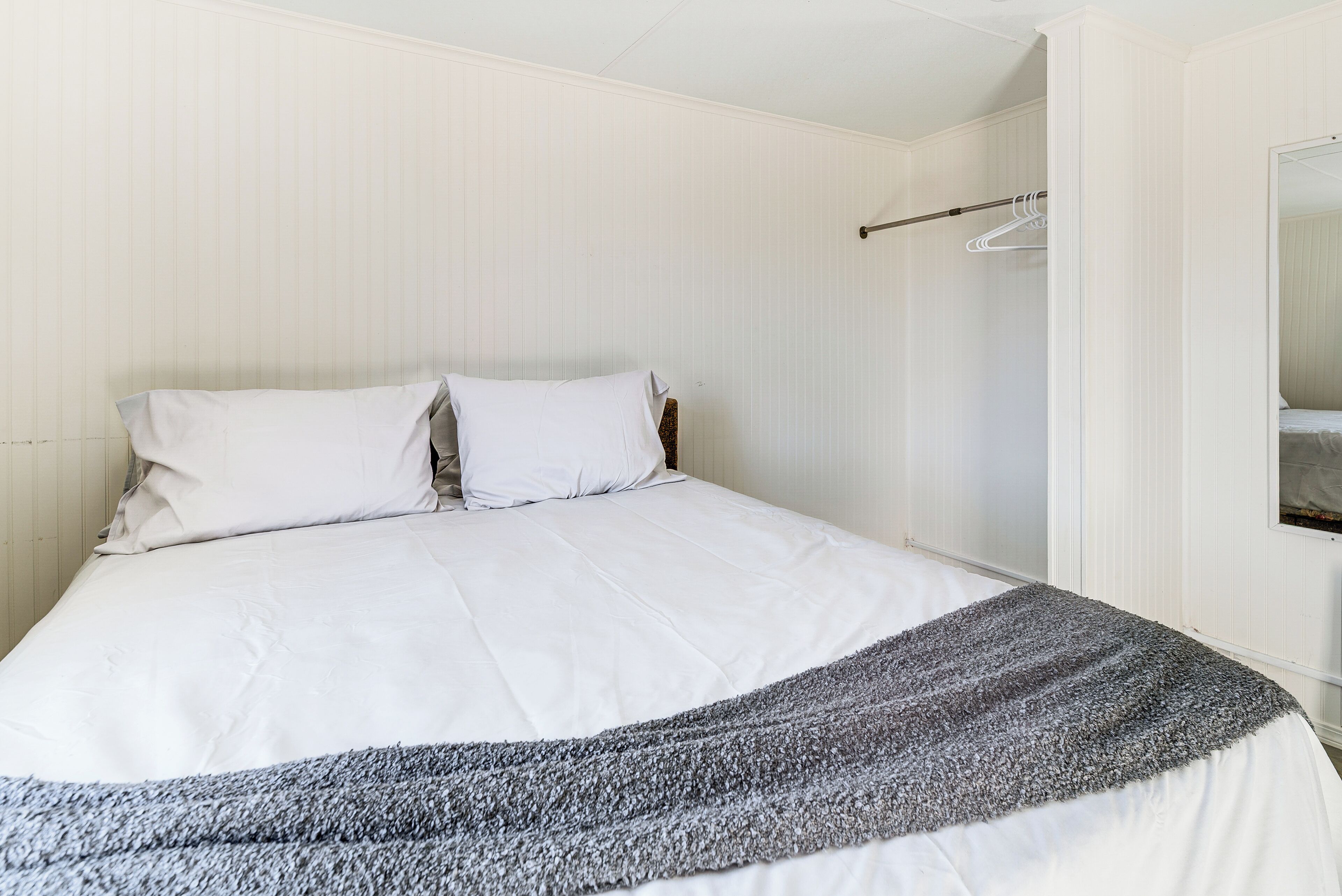 A neatly made bed with white linens and a gray blanket, a wooden headboard, and a white wall with a hanging coat rack.