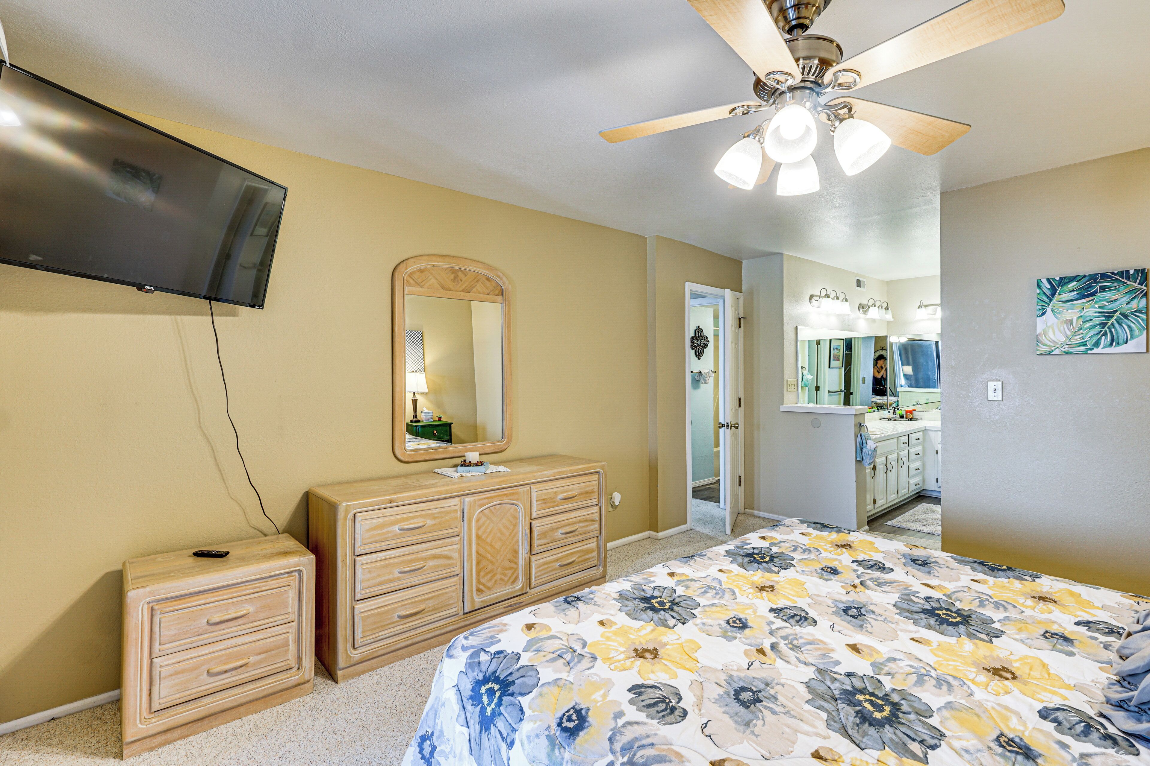 A bedroom with a bed, a dresser, a mirror, a ceiling fan, and a TV mounted on the wall.