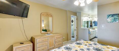 A bedroom with a bed, a dresser, a mirror, a ceiling fan, and a TV mounted on the wall.