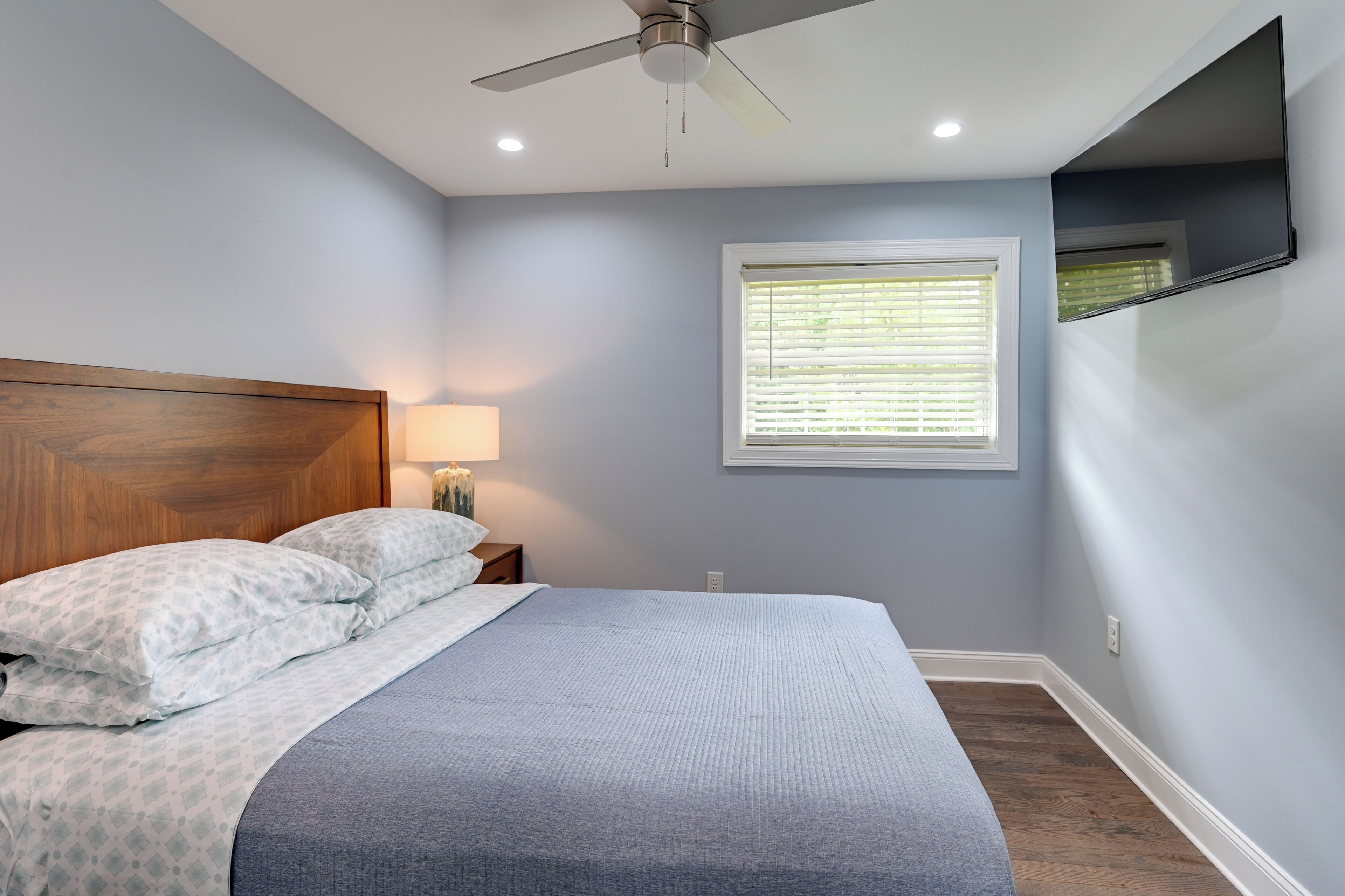 Une chambre à coucher comprenant un lit, un ventilateur de plafond, une table de chevet, une lampe, une fenêtre avec des stores et un téléviseur fixé au mur.