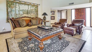 A living room with a sofa, a leather ottoman, a chair, a large tapestry, and a lamp. - Golf, Swim & Grill: Snowbirds Escape in Sun Lakes (Chandler)