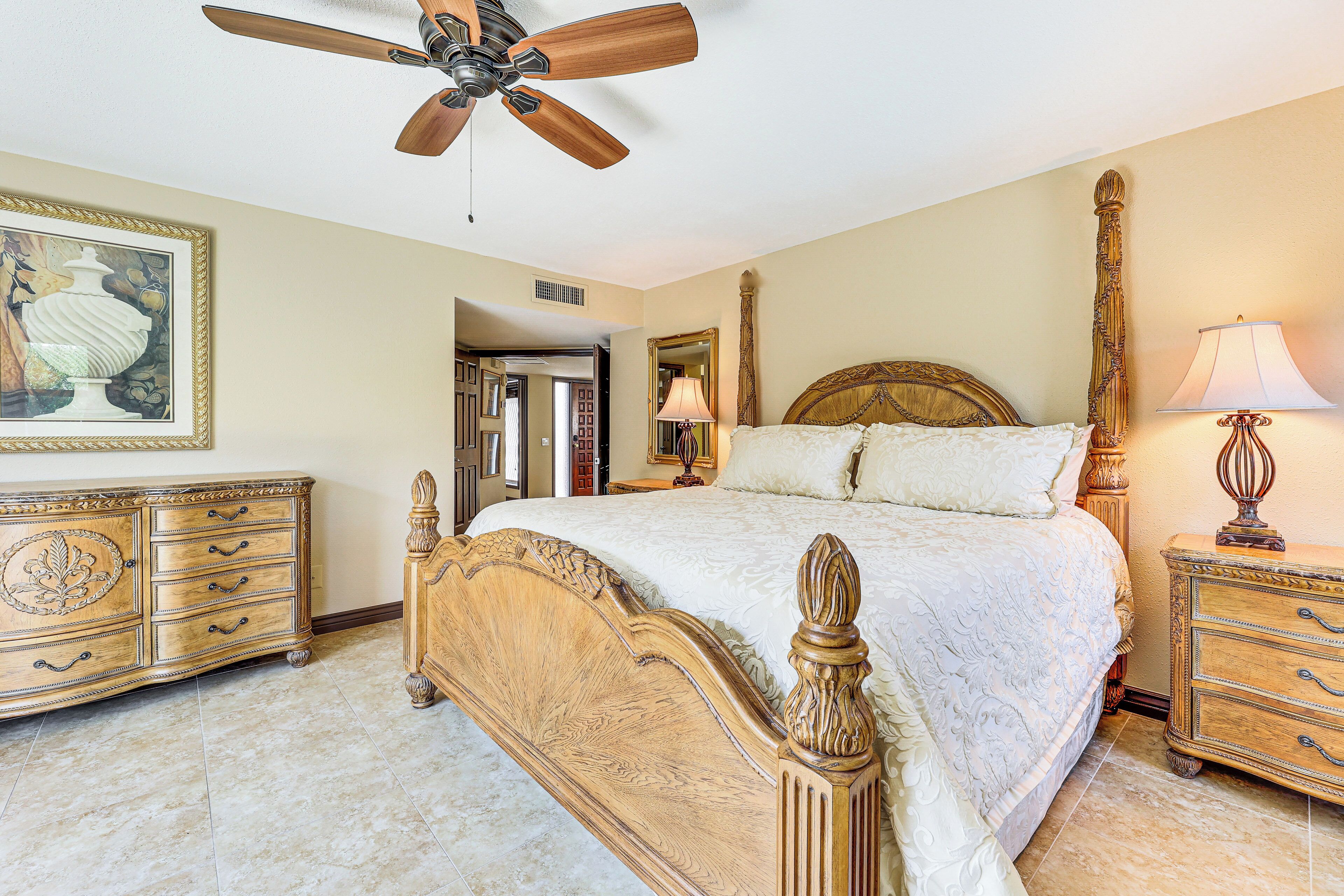 A bedroom with a large bed, wooden headboard, a ceiling fan, a dresser, and a framed picture on the wall.