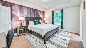 A bedroom with a bed, two bedside tables with lamps, a chair, a window with curtains, and a wall with a forest mural.