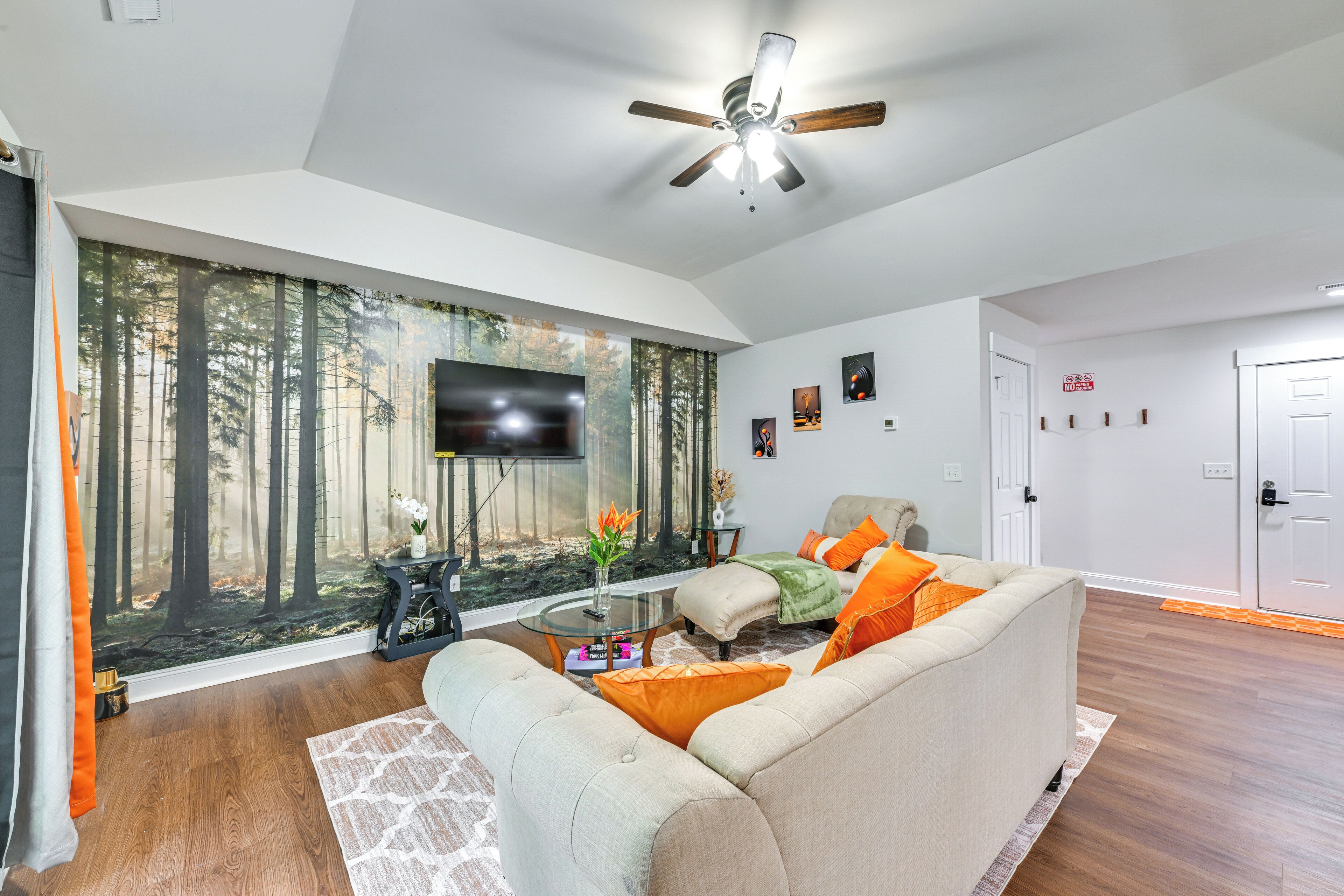 Un salon moderne doté d’une grande fenêtre, d’un ventilateur de plafond, d’un téléviseur à écran plat et d’un canapé confortable agrémenté de coussins orange.