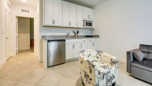 A modern kitchen with white cabinets, a stainless steel dishwasher, and a microwave. There is a patterned armchair and a gray sofa in the living area. - Beach-hop & Golf! Port St Lucie Apt w/ Shared Yard (Port Saint Lucie)