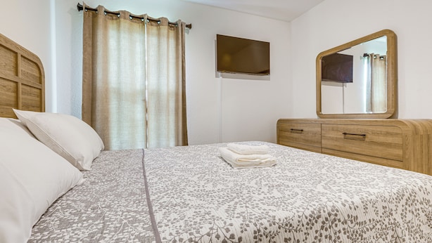 A neatly made bed with a floral-patterned bedspread, a wooden headboard, a bedside table with a mirror, and a window with curtains. - Beach-hop & Golf! Port St Lucie Apt w/ Shared Yard (Port Saint Lucie)