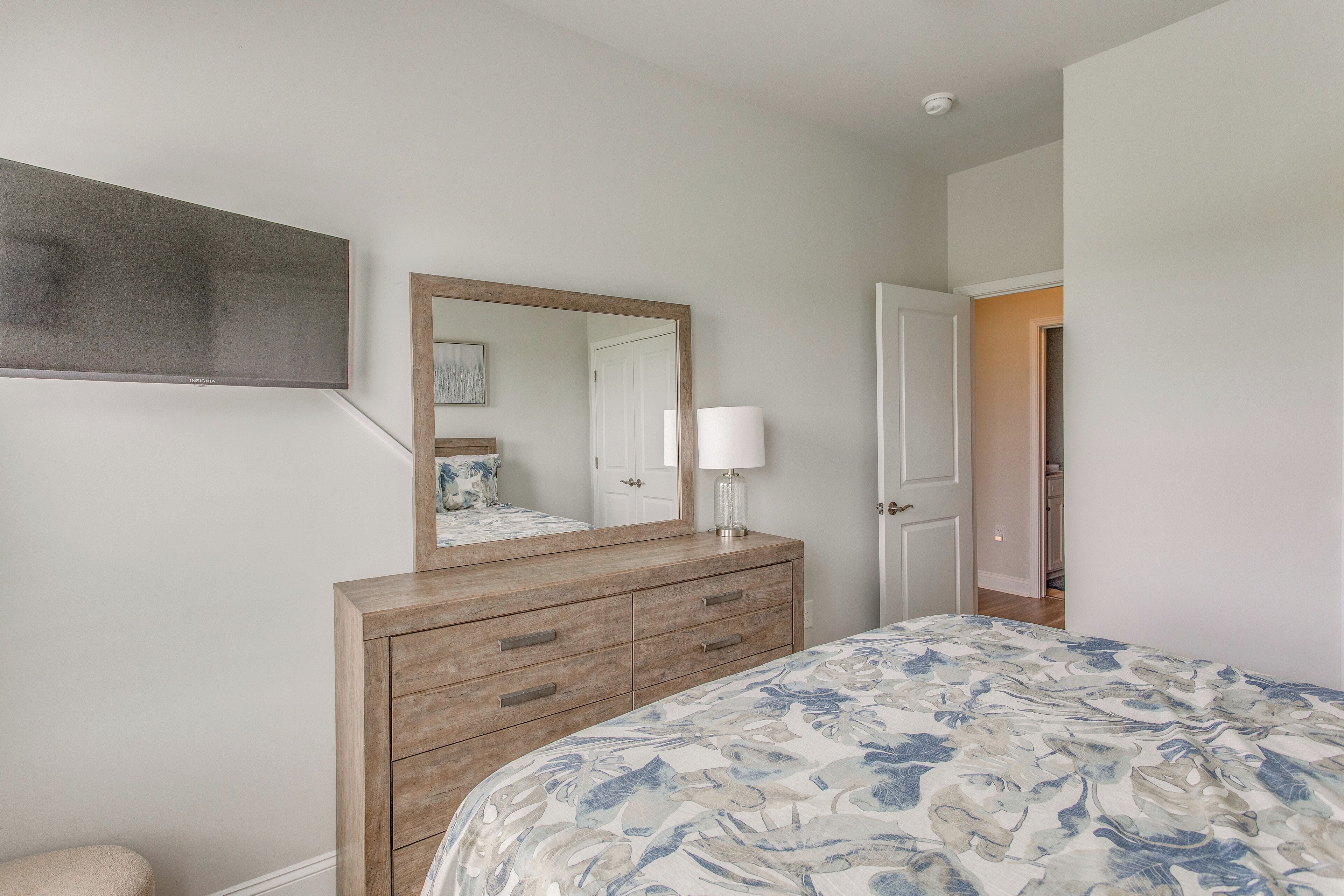 A bedroom with a large bed, a wooden dresser, a mirror, and a door leading to another room.