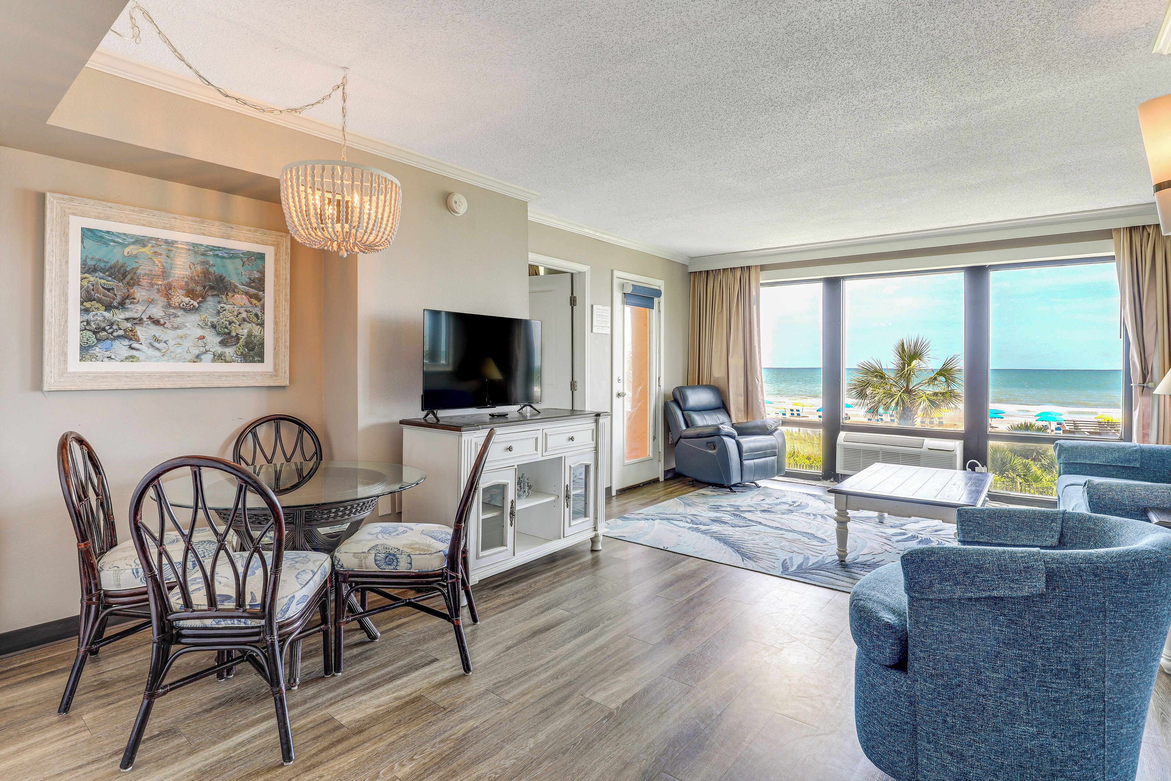 A hotel room with a dining area, a TV, a sofa, and a view of the ocean.