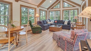 A spacious living room with wooden floors, large windows, and a high ceiling with exposed beams. There is a dining table with chairs and a sofa set. - Waterfront Escape on Big Sand Lake w/ Private Dock (Minong)