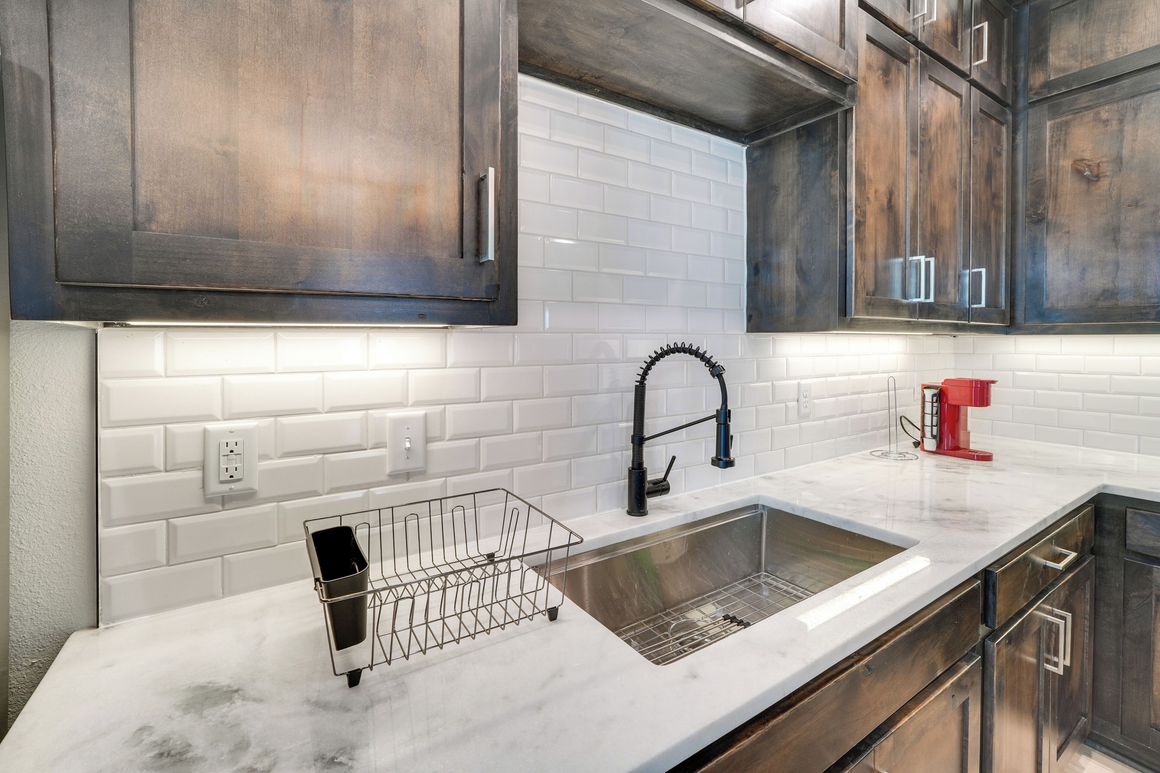 A modern kitchen with dark wooden cabinets, white subway tiles, a stainless steel sink, and a red coffee maker.