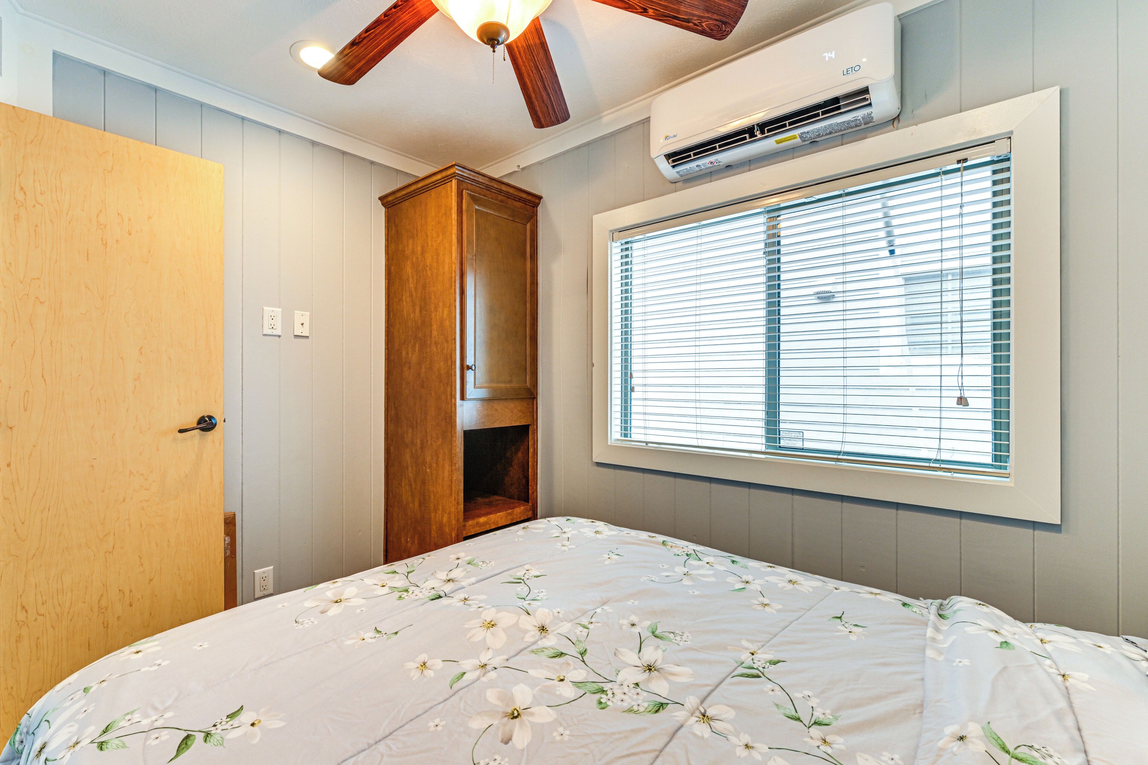 A bedroom with a bed, a wooden wardrobe, a window with blinds, and an air conditioning unit.