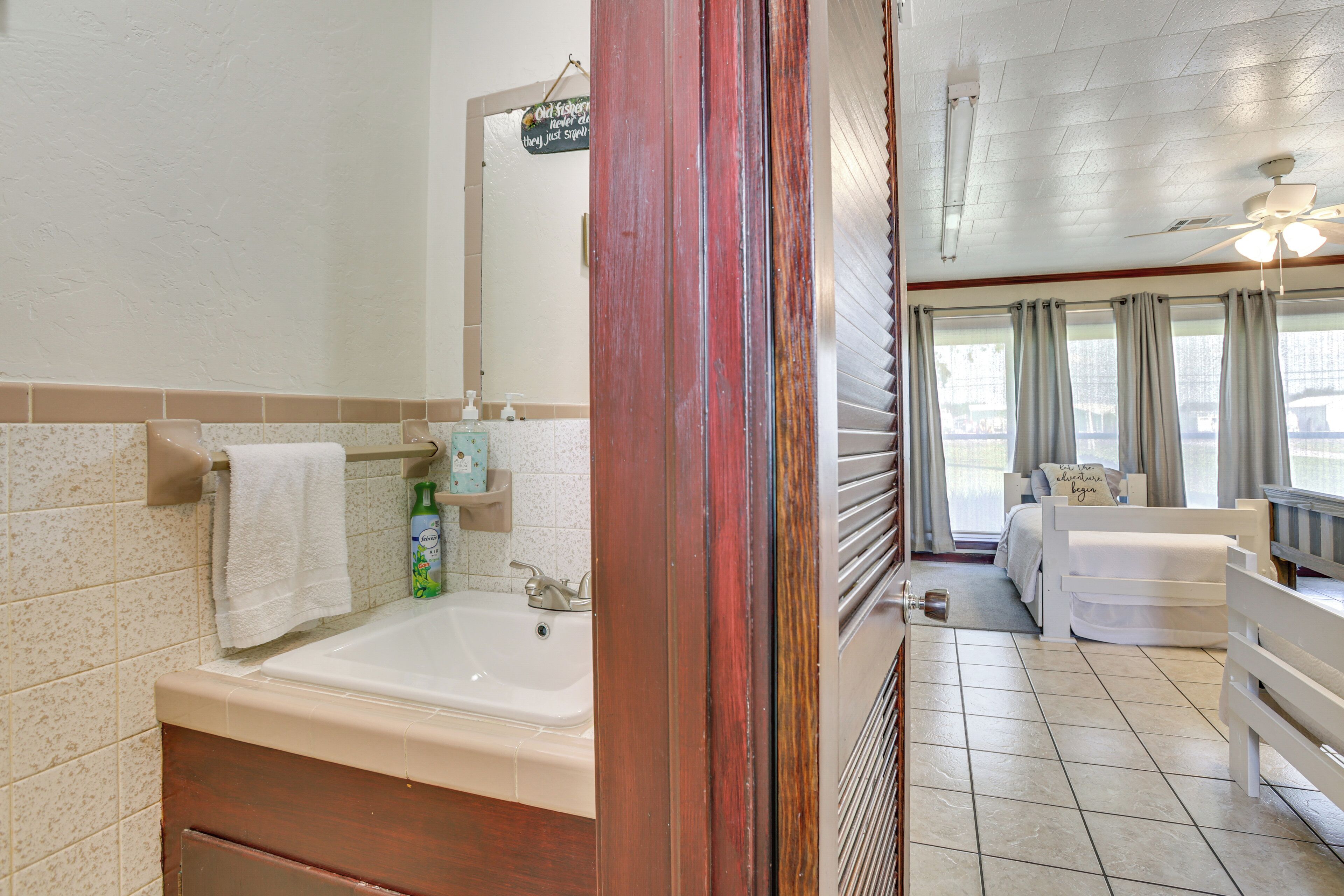 Une salle de bain avec une baignoire, un lavabo et un miroir. Il y a une fenêtre avec des rideaux et, au fond, un lit avec une tête de lit blanche.