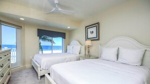 Een hotelkamer met twee bedden, een dressoir, een plafondventilator, een ingelijst schilderij aan de muur en uitzicht op de oceaan door het raam.