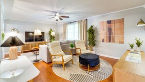 Un salon moderne comprenant un canapé, deux fauteuils, une table basse, un ventilateur de plafond, un téléviseur et une petite table d’appoint avec une lampe.