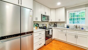 Une cuisine moderne avec des armoires blanches, des appareils en acier inoxydable, une fenêtre donnant sur un espace verdoyant et un plancher en bois.