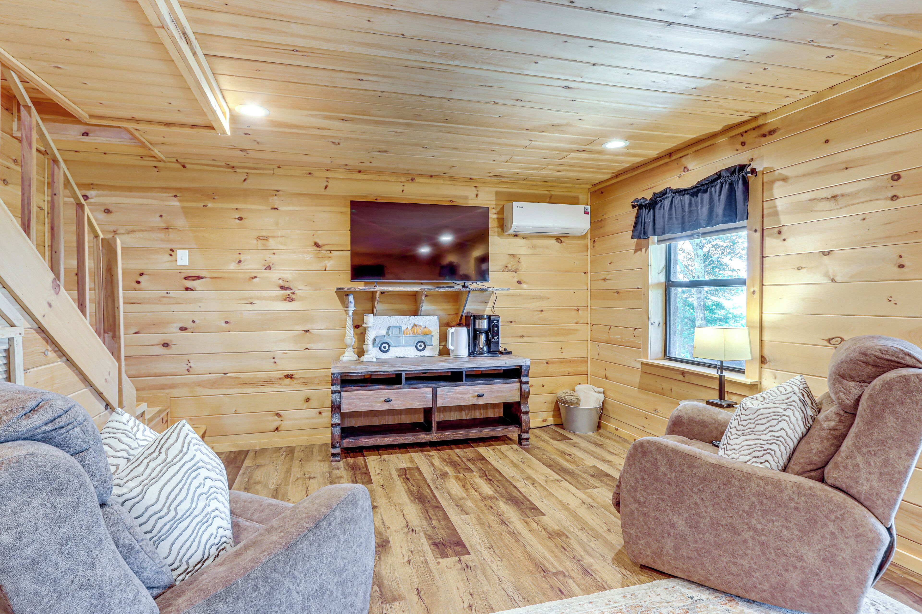 A cozy cabin interior with wooden walls, a sofa, a recliner, a wooden entertainment unit, and a window with a view of trees.