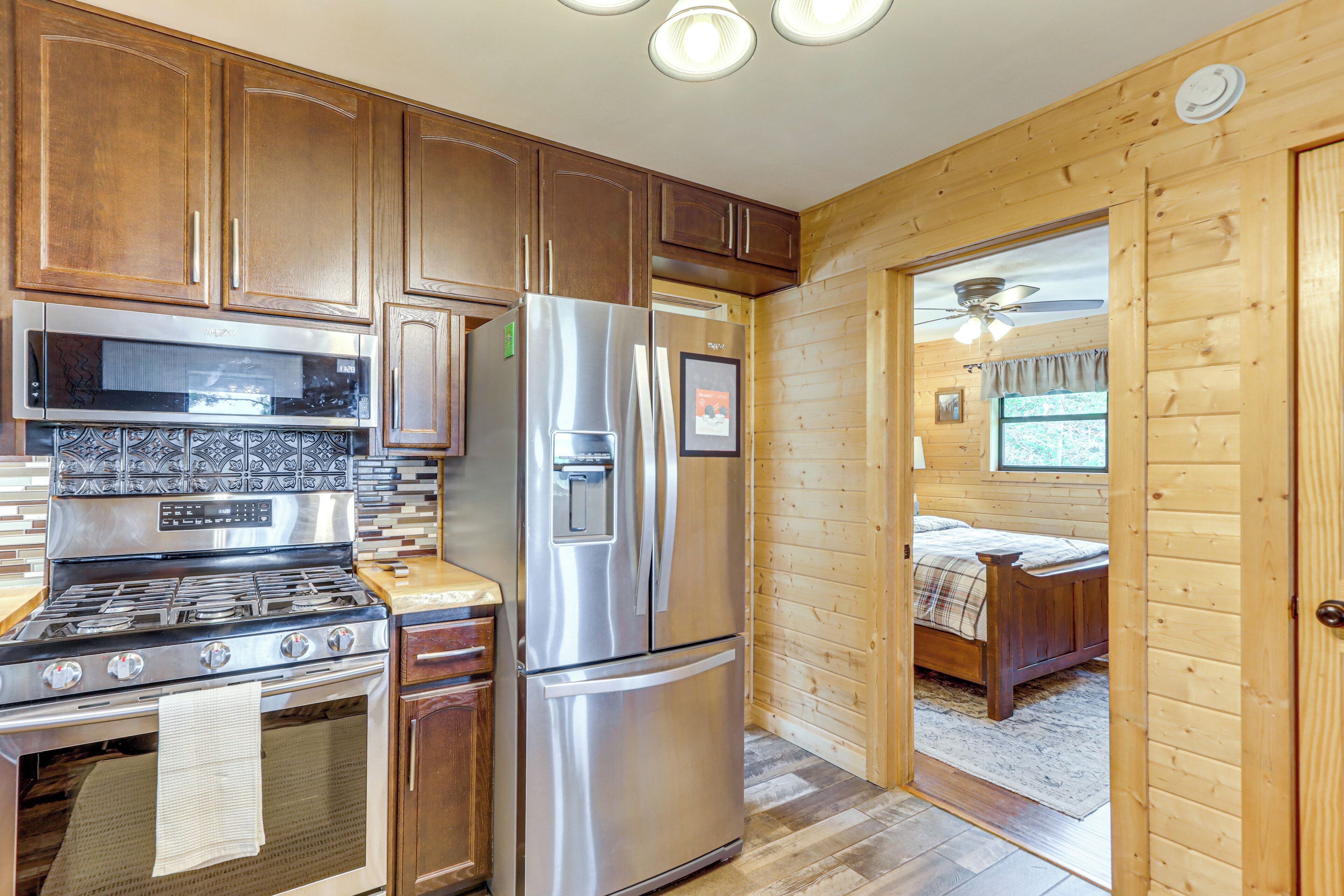 A compact kitchen with stainless steel appliances, wooden cabinets, and a visible door leading to a bedroom with a bed and a ceiling fan.