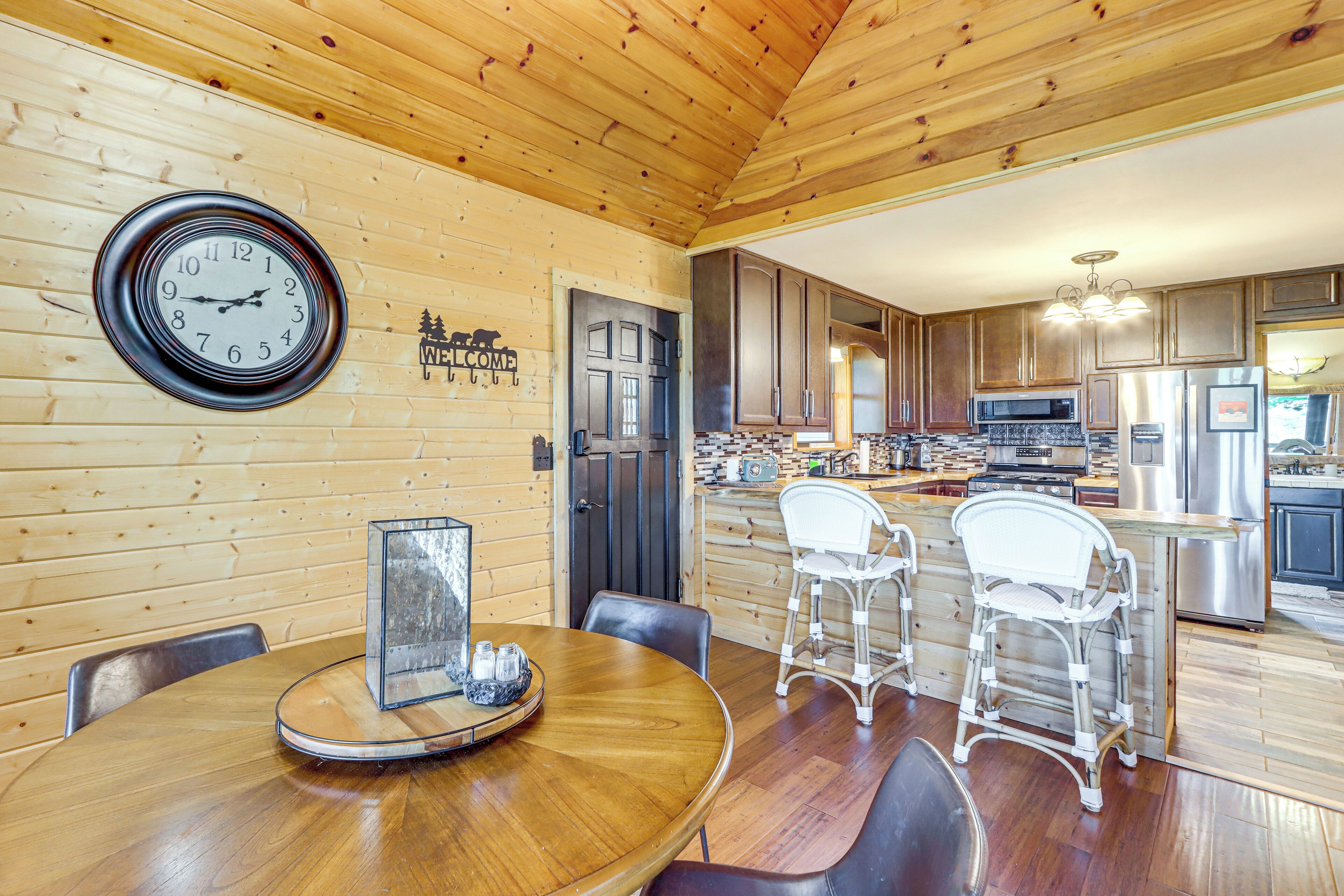 A wooden interior with a dining area featuring a round table and chairs, a kitchen with modern appliances, and a clock on the wall.