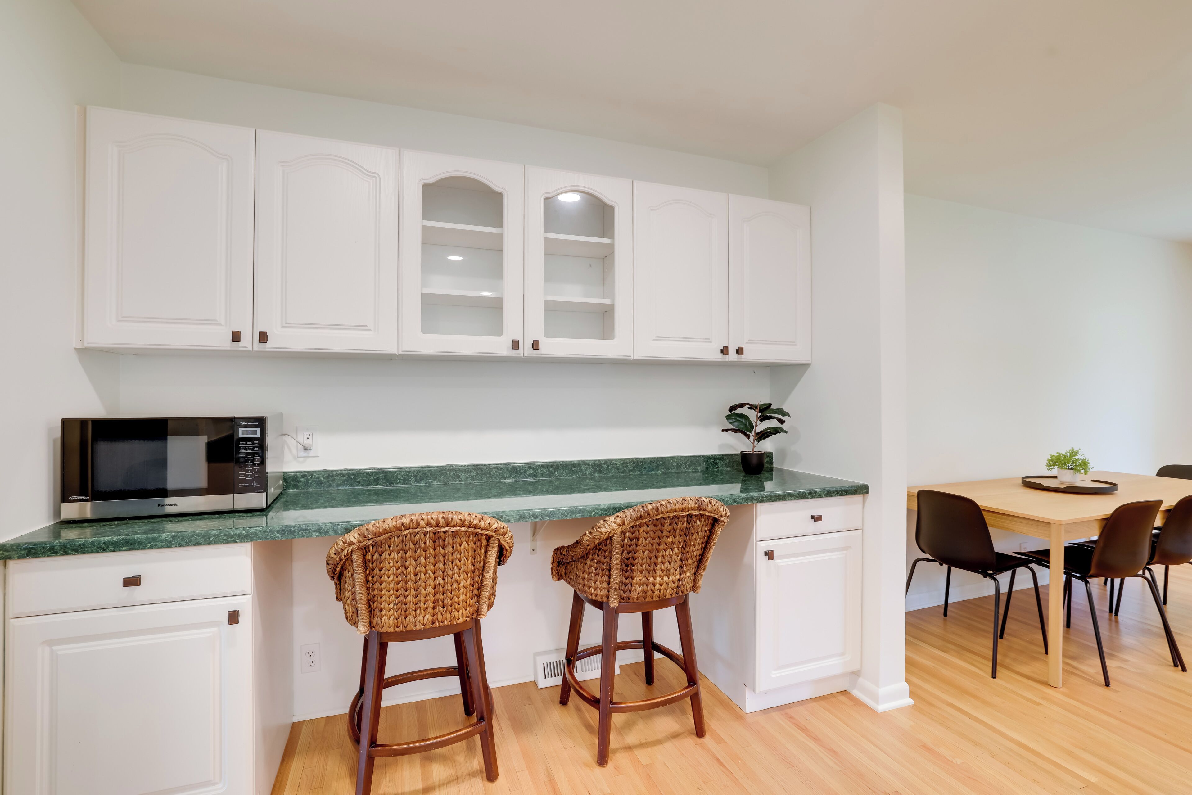 A kitchen with white cabinets, a microwave, two wicker bar stools, a dining table with chairs, and a potted plant.