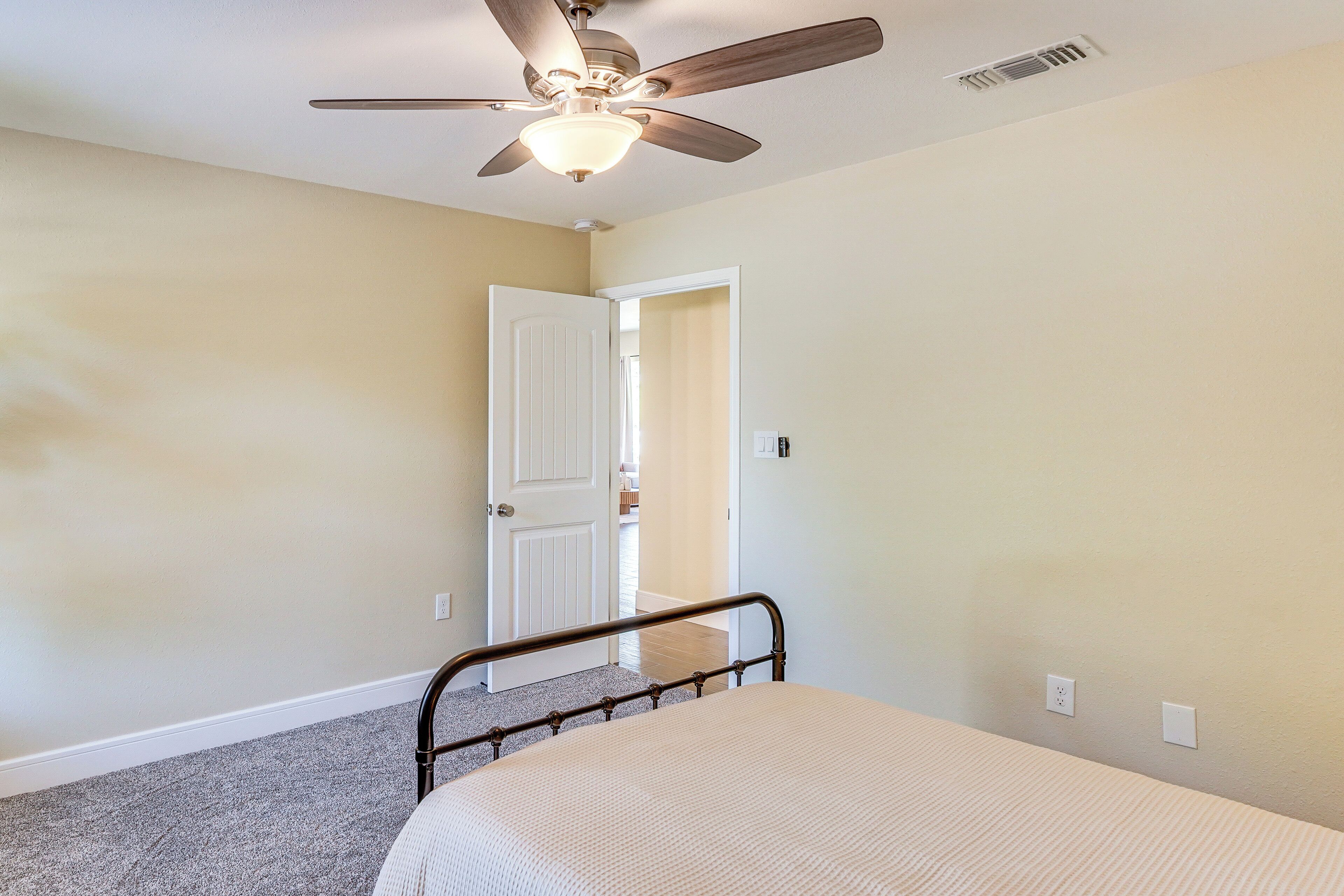 A bedroom with a bed, a ceiling fan, a door leading to another room, and a carpeted floor.