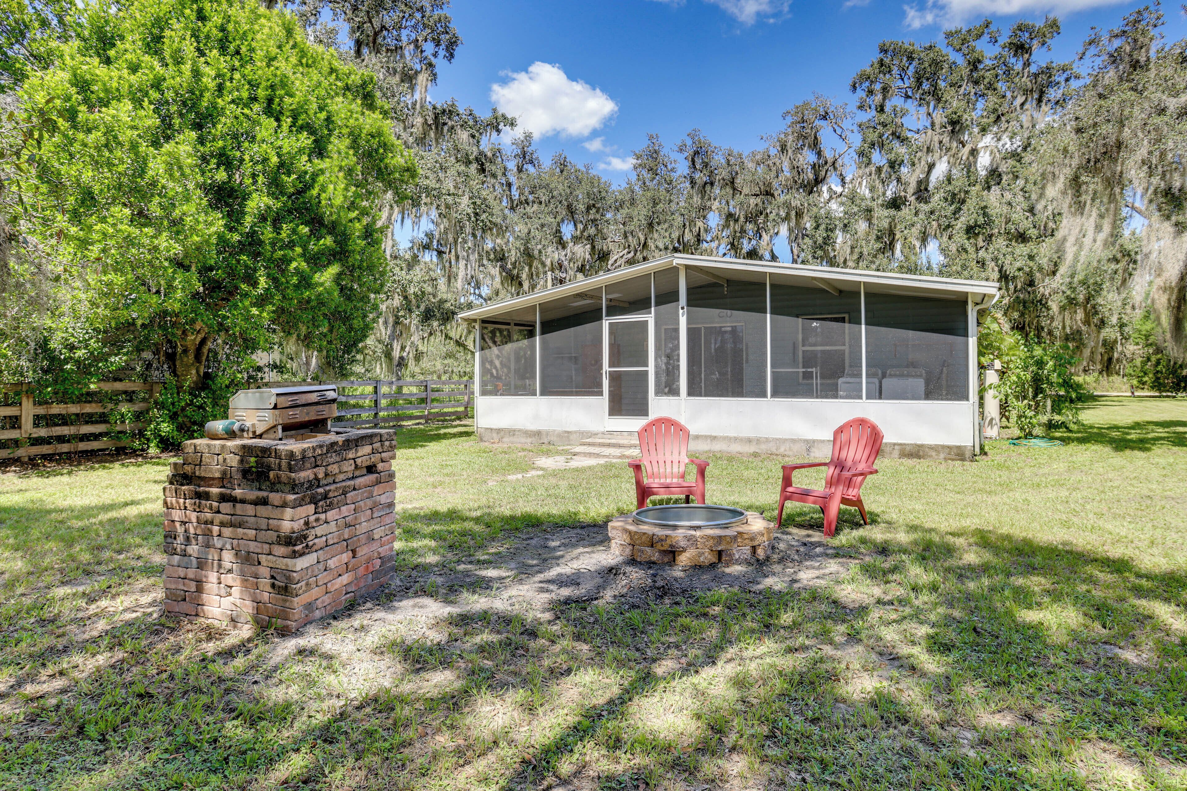 A covered outdoor seating area with two red Adirondack chairs, a brick fire pit, and a view of a white building with a screened porch.