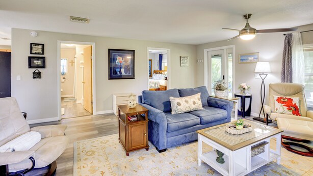 Un salon avec un canapé bleu, un fauteuil blanc et une table basse. On y trouve une petite table d’appoint en bois et un lampadaire. La pièce est équipée d’un ventilateur de plafond et d’une fenêtre avec des rideaux.