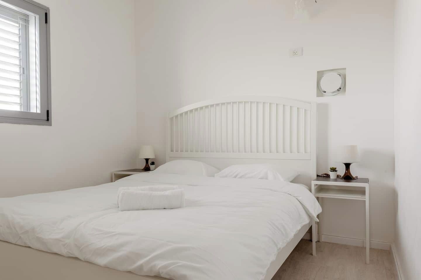 A white bedroom with a bed, two bedside tables, and a window.