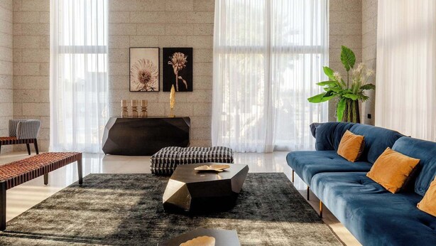 Una sala de estar moderna con un sofá, una mesa de centro y una mesita auxiliar. Hay un ventanal con cortinas traslúcidas, un estante montado en la pared con objetos decorativos y una planta en maceta.