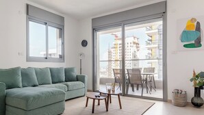 Una sala de estar moderna con un sofá verde, una mesita redonda y un gran ventanal corredero que da a un balcón al aire libre con vistas a la ciudad.