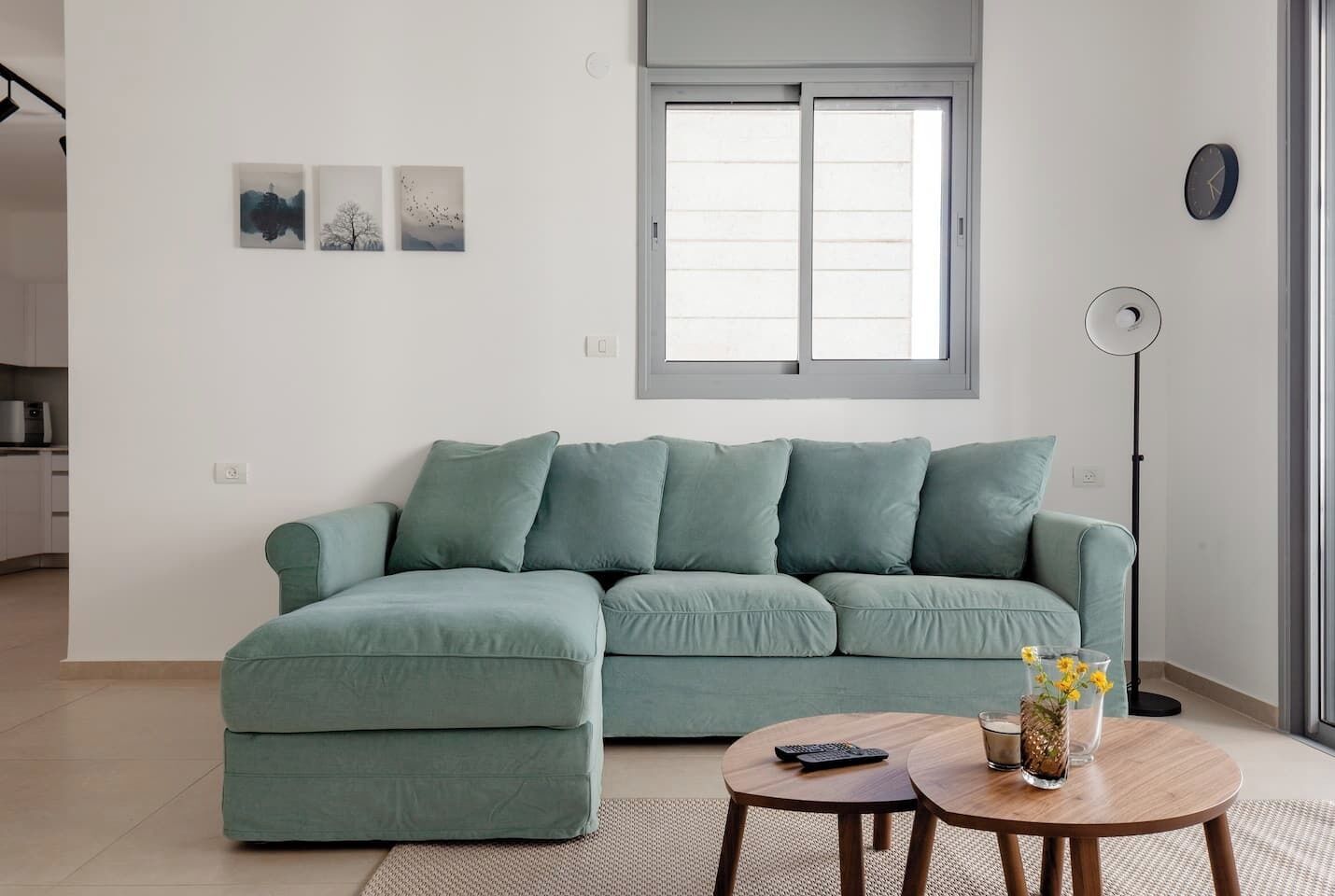 Un salón con un sofá verde, una mesa de centro redonda de madera y una lámpara de pie. Hay tres cuadros enmarcados en la pared y una ventana con persianas.