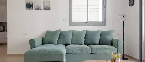 Un salón con un sofá verde, una mesa de centro redonda de madera y una lámpara de pie. Hay tres cuadros enmarcados en la pared y una ventana con persianas.