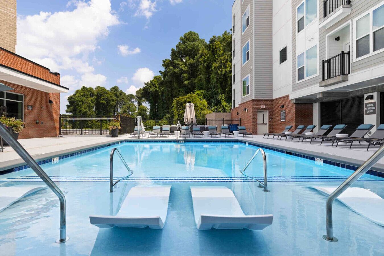 A swimming pool with lounge chairs and umbrellas, surrounded by buildings and trees.