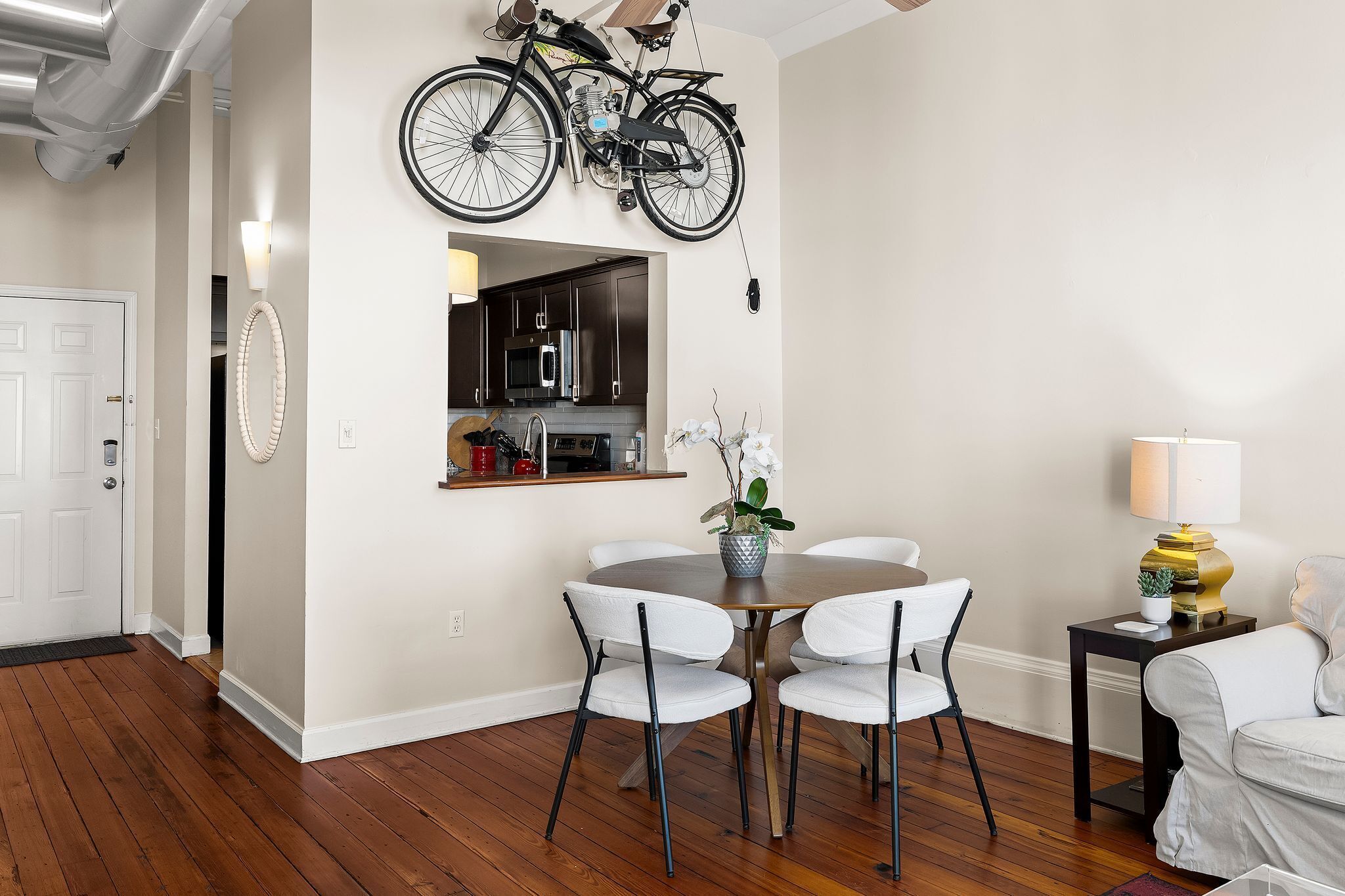 A dining area with a round table, four white chairs, a potted plant, and a bicycle mounted on the wall.