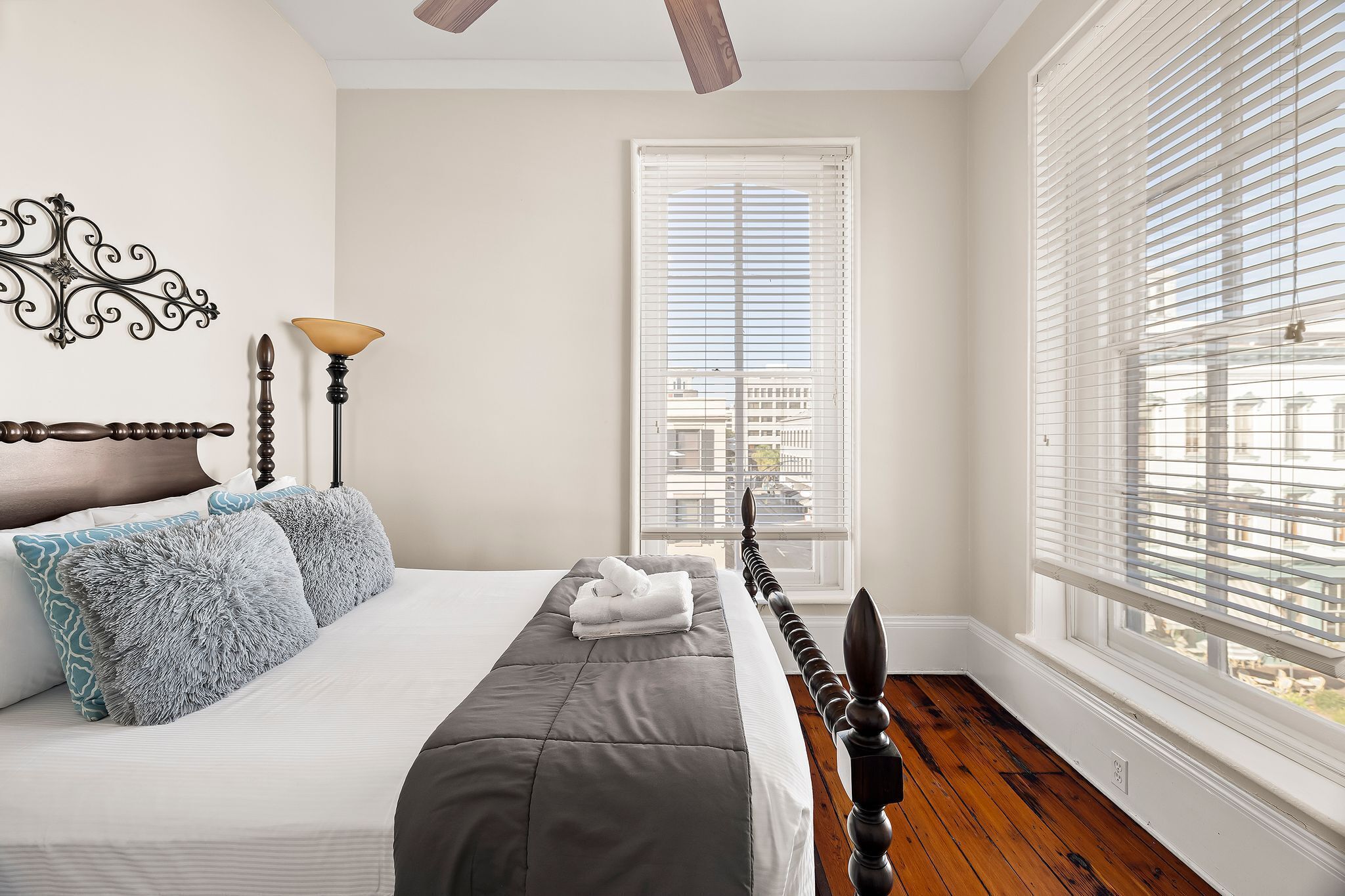 Un lit bien fait, recouvert d’une couvre-lit grise et agrémenté de coussins décoratifs, un plancher en bois, un ventilateur de plafond et une fenêtre avec des stores.