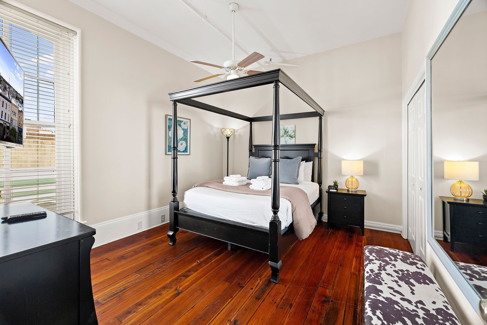 A four-poster bed with a ceiling fan, bedside lamps, and a view of buildings through the window.