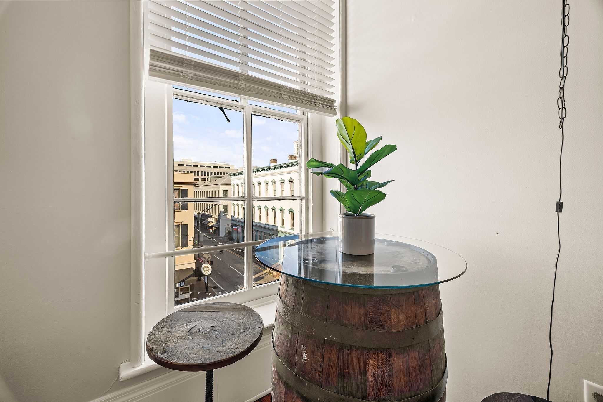 Um quarto com uma grande janela, uma mesa redonda de madeira, um pequeno banquinho e uma planta em vaso.
