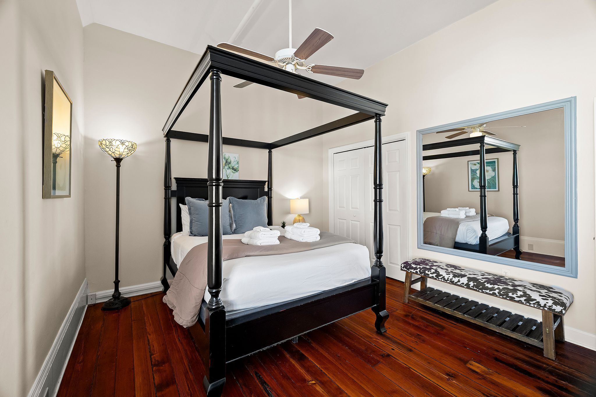 A four-poster bed with a ceiling fan, a bench, and a large mirror in a bedroom with hardwood floors and white walls.